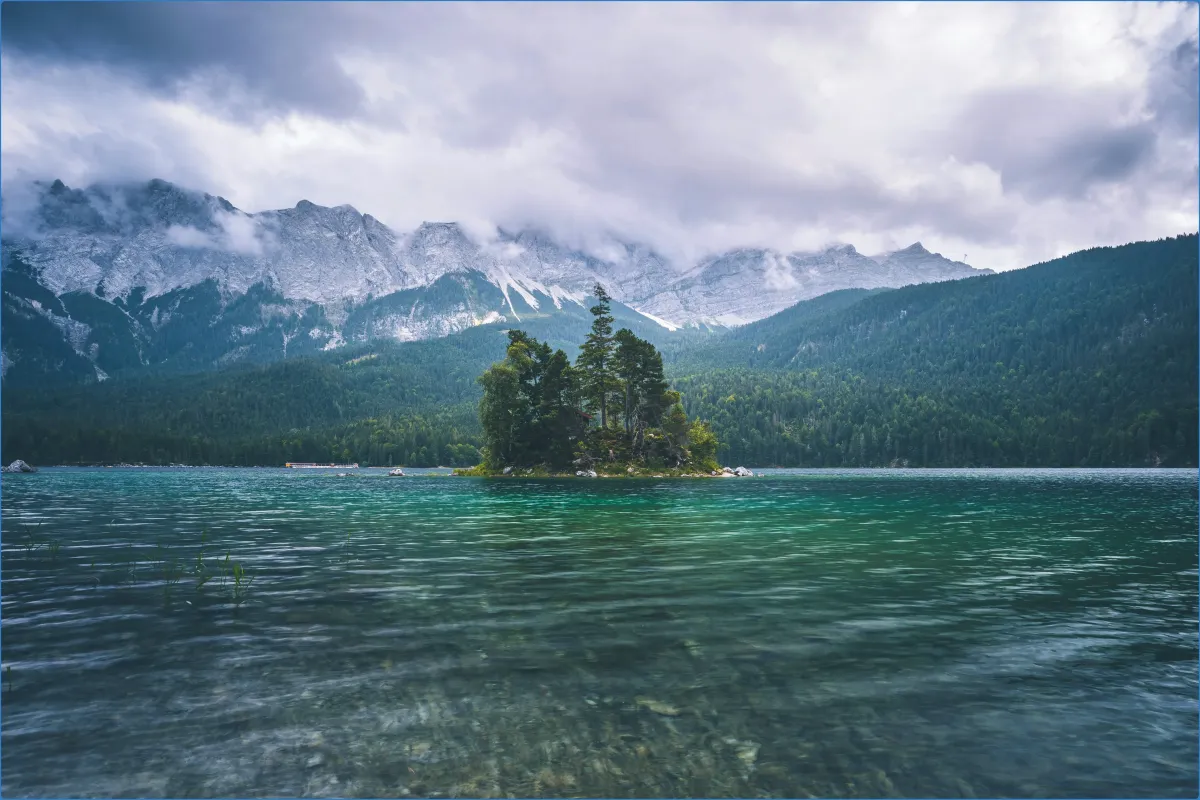 Kleiner bewaldeter Insel im klaren See vor Bergkulisse.