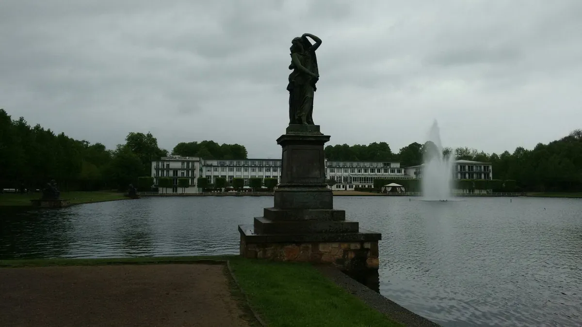 Statue vor einem Teich mit Springbrunnen und Gebäude im Hintergrund.