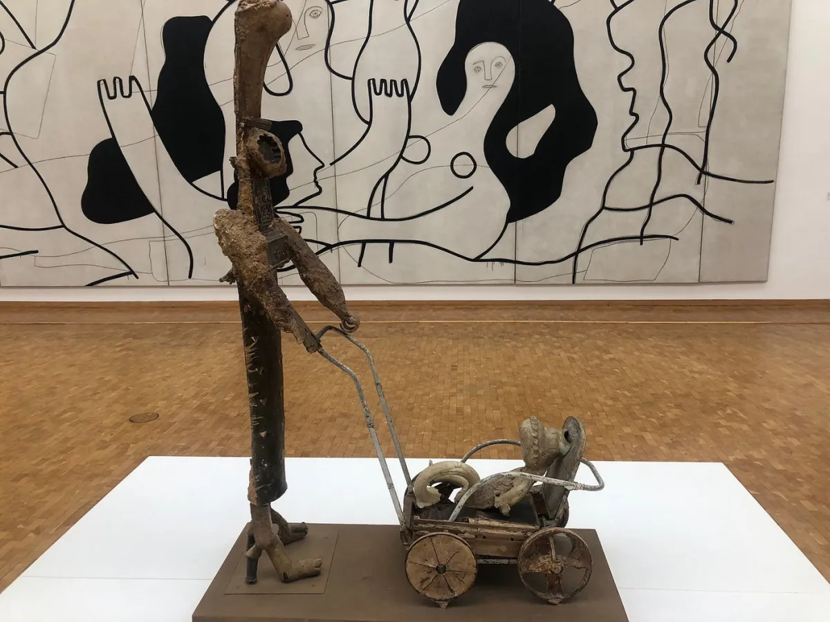 Skulptur eines Menschen mit Kinderwagen vor einem Gemälde.