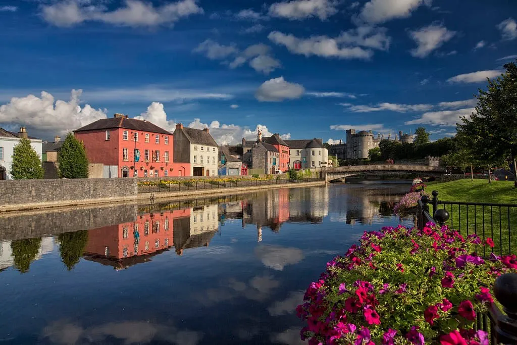 Colorful buildings and flowers by a river with a bridge.