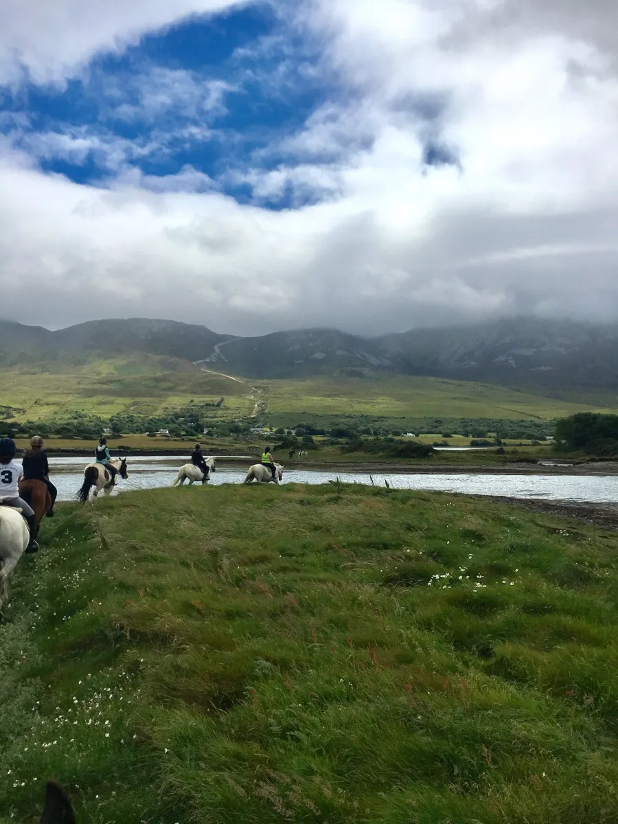 People horseback riding near a river with mountains in the background.