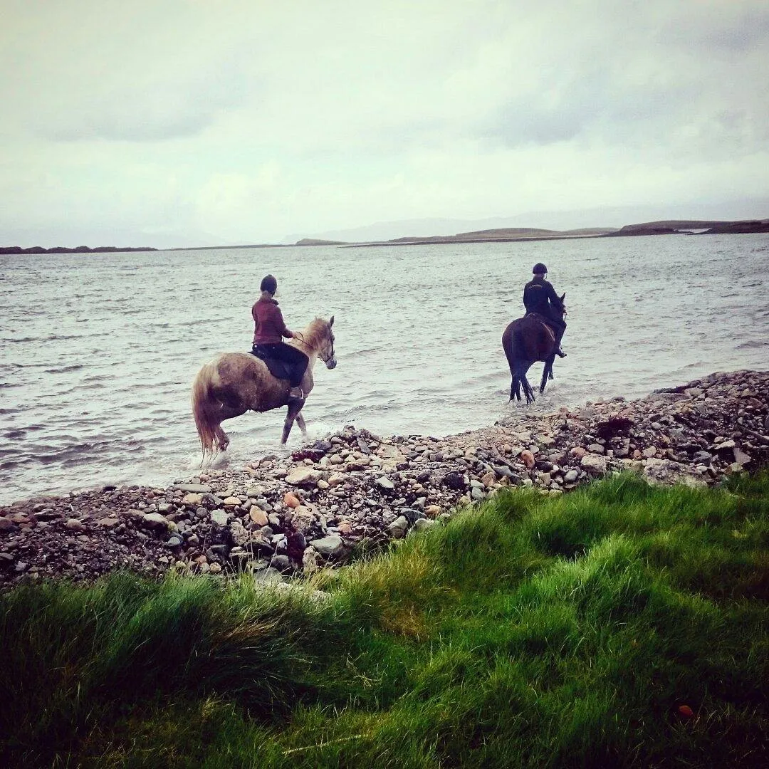 Two people horseback riding along a rocky shoreline.