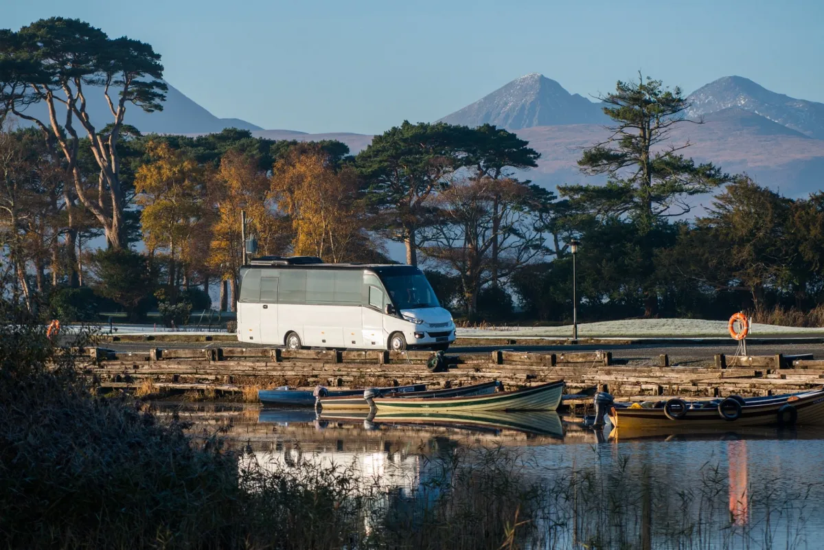White bus parked near a lake with mountains in the background.