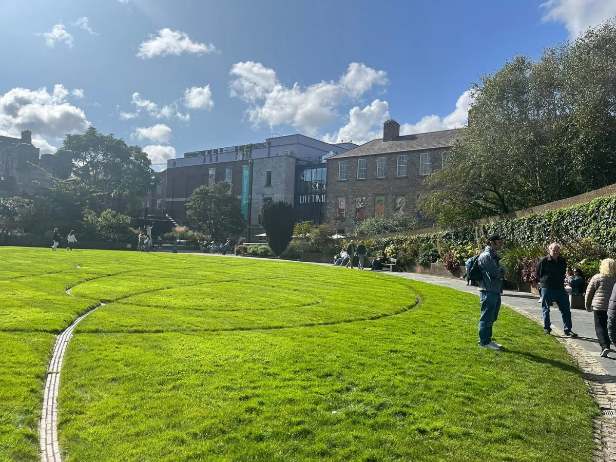 People walking and sitting in a sunny park with buildings nearby.