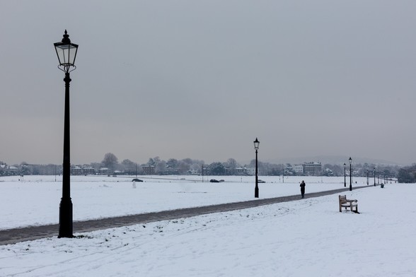 A snowy open space with a path punctuated by old-fashioned lampposts receding from left to right across the frame.