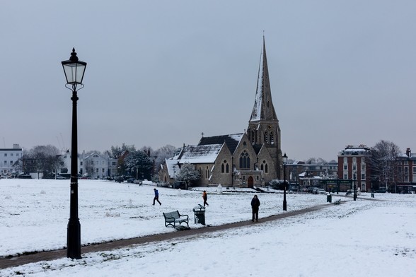 A snowy open space with a path punctuated by old-fashioned lampposts receding from left to right across the frame, with a church and spire in the middle.