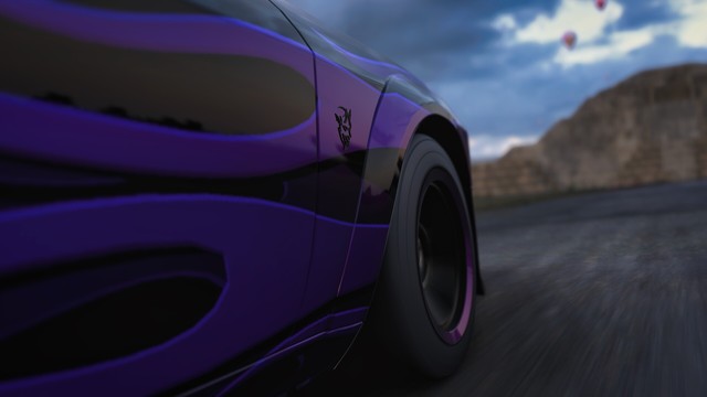 Capture of the front quarter panel, tire, and Demon logo of a black Dodge Challenger SRT Demon with purple two-tone flames from the front at ground level on a gravel road.
