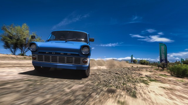 Front-facing capture of a blue 1965 Ford Transit van with a Vault-Tec emblem from Fallout drifting sideways from the right of the frame to the left on a dirt trail with cactus, leafy trees, and blue skies in the background. Blue drift zone banners appear along the path.