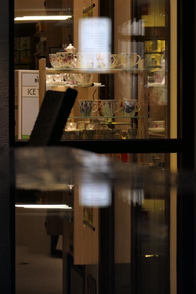 Ein erleuchtetes Schaufenster bei Nacht, in dem Porzellangeschirr (hauptsächlich verschieden bemalte Tassen in unterschiedlichen Formen) ausgestellt ist. Im Laden sieht man außerdem noch einen Teil einer Truhe, aus der wahrscheinlich Eis verkauft wird (man sieht den Schriftzug "ICE" darauf). In der Scheibe spieget sich ein weißes Blatt Papier oder Schild mit unleserlicher Aufschrift. Das Ganze spiegelt sich in einer Pfütze auf einem Tisch.  
An illuminated shop window at night, displaying porcelain tableware (mainly various painted cups in different shapes). In the store you can also see a part of a chest from which probably ice cream is sold (you can see the lettering "ICE" on it). In the window there is a white sheet of paper or a sign with an illegible inscription. The whole thing is reflected in a puddle on a table.