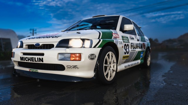 Closeup of 1992 Escort RS Cosworth taken from the front passenger size after a heavy rain. Water beads on the retro white, forest green, and gray Fujifilm livery and puddles reflect the car and sky above.