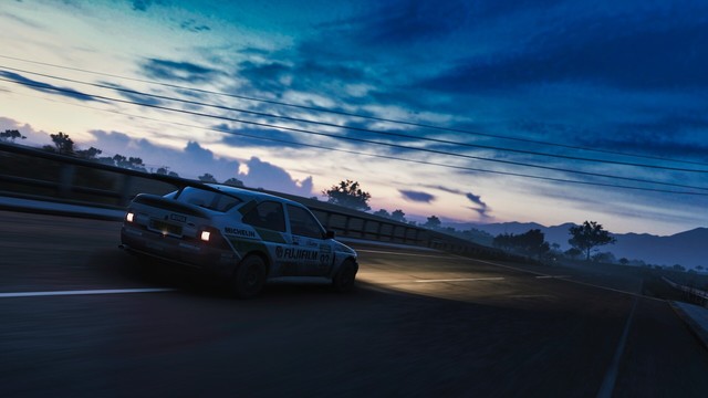 Following shot of 1992 Escort RS Cosworth in a retro green, gray, and white Fujifilm livery taken from the rear passenger side near sunrise. Thunderheads line the horizon and the sky fades from light pink to dark blue.