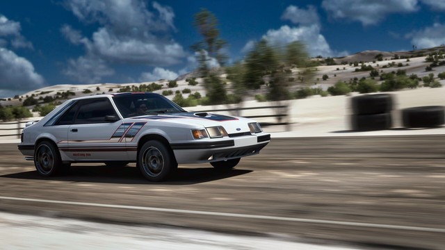 Moving shot of a 1986 Ford Mustang SVO moving from left to right in the frame on a paved road throughs sand dunes. Blue skies with thunderheads loom in the distance.