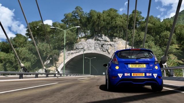 Shot from behind of a 2014 Ford Fiesta ST hatchback as it nears a tunnel at the end of a suspension bridge. Trees line the rocky tunnel entrance and lights trail off in the tunnel to darkness.