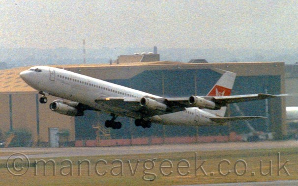 Side view of a large white 4 engined jet airliner taking off from an airport, with the undercarriage still extended.
There are no markings on the body, but there is a thick red stripe with a winged circle on the tail.
Behind it is a large brown hangar, above which the sky is a dreary grey.