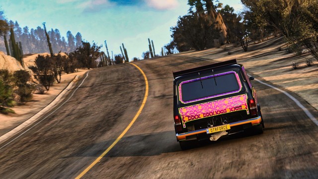 Shot from behind of a 1994 Ford Supervan 4 in a patterned black, orange, purple, and gold livery reminiscent of an ornate rug. The van is speeding up a hill and around a bend through a rocky gap. Cacti and brush line the road. The golden hues of sunset tint the photo.