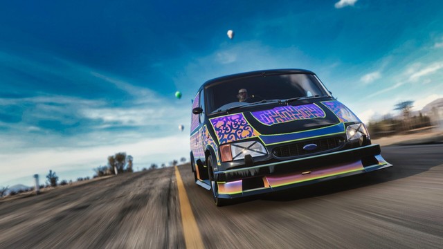 Close up shot from the front of a 1994 Ford Supervan 4 in a patterned black, orange, purple, and gold livery reminiscent of an ornate rug.  The van is cresting  a hill and the wispy clouds in the sky form a crown, moving away from the van in all directions. The driver focuses on the road intensely. He is wearing a red clown nose.