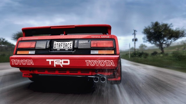 Close up shot from the rear of a 1989 Toyota MR2 SC with a red TRD livery and TRD and Toyota badges on the bumper. The road is wet and water streams off the tires as storm clouds darken the horizon.