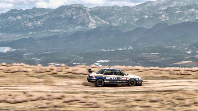 Distance shot from the righthand side of a 1990 Subaru Legacy RS with a bodykit, aero, and Foster's beer livery speeding down a dirt road on the edge of the volcano. The shot shows most of the valley in the distance, including the ruins of an ancient Mexican temple, agricultural fields, and two small villages. Mountains loom even further in the distance.