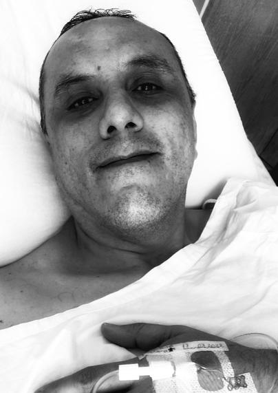 Un hombre en una cama de hospital, con una sonda intravenosa visible en el dorso de su mano. Su rostro se ve agotado pero parece cerca de sonreír.