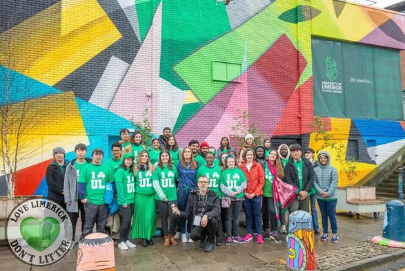 A photograph of a large group of University of Limerick students in green t-shirts which say "I heart UL", with the heart emoji replaced by a shamrock. They are stood next to a brightly coloured building, with a painted mural made of geometric shapes. Credit: I Love Limerick