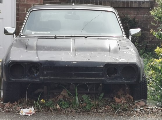 Image shows the front of a black Reliant Scimitar GTE parked in the front garden of a house. The car is missing the front grille and headlamps but appears to otherwise complete.