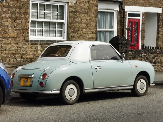 A Nissan Figaro with a white roof over a pale green body. The car is parked in a suburban street.