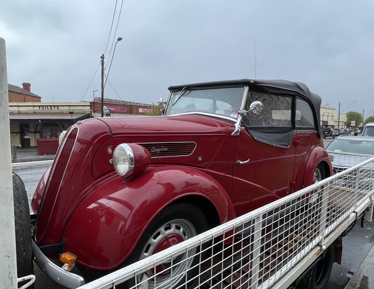 A red 1950s Ford Anglia convertible on a trailer. The word Anglia is visible on the side of car above the fender.