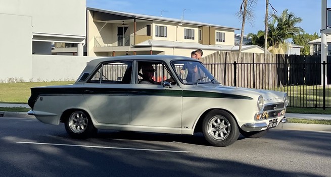 A 1960's Ford Lotus Cortina MK1 4 door salon. The car is white with a signature green stripe that runs from  the rear lights to the front wing. The stripe narrows ending in a point just short of the headlamp. The car is on period Roystle wheels and may have been slightly lowered. 
The car is driving along an Australian street watched by some young boys.