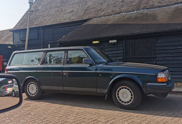 Side view of a dark blue, looks sort of green in the photo, Volvo 240 SE Estate. The car is parked in front of a historic looking black timber barn like building.