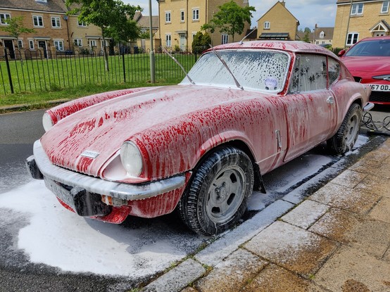 A red car - a 1972 Triumph GT6 Mk3 - sitting on the road covered in white 'snow foam'. Some houses and an area of grass are visible behind it.