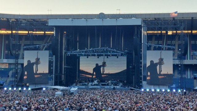 Bruce Springsteen holding up his guitar in one hand in front of the sunset, on three giant screens on the stage at Murrayfield Stadium
