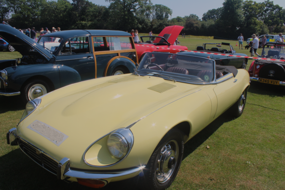 Front 3/4 view of a Jaguar E-Type convertible finished in Primrose Yellow. The sun is shining, and the car is parked with it roof lowered.