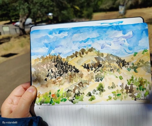 Watercolor sketch of California hillside in sketchbook. Blue sky, golden brown hills with dark trees, green in foreground.