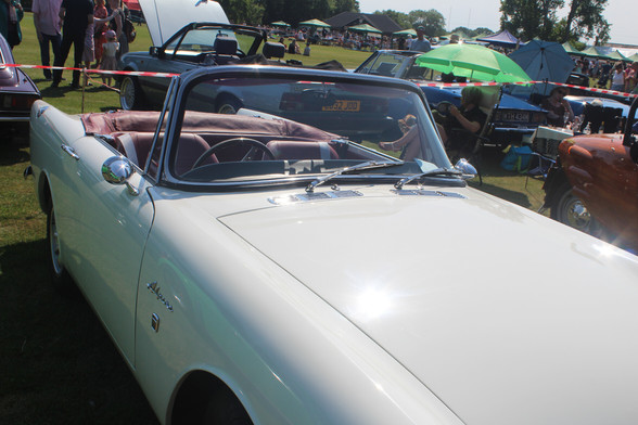 Front 3/4 view of a white 1969 Sunbeam Alpine convertible, the name "Alpine" is clearly visible on the wing behind the wheel arch above a 1750 badge. It is  sunny day and the roof is revealing a red interior.