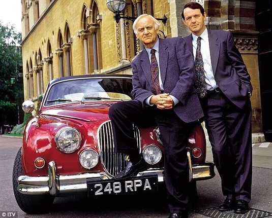 A promotion image from the TV show Inspector Morse. The titular main character is leaning against the front of a red Jaguar MKII with his weight on the front of the left wing. His assistant Sargent Lewis is stood by the side of the car with his right hand on the left wing. Morse is played by John Thaw and Lewis by Kevin Wheatley.