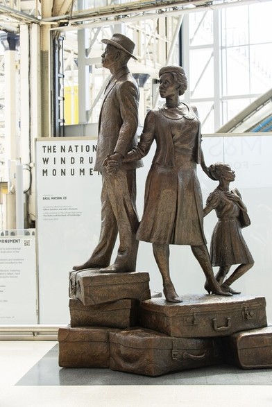 A family group of a father, mother, and child stand atop a pile of suitcases. The are all linked by touch. The following description is from Basil Watson, the sculptor: 

"Then within the composition of the figures, there are elements of the man and woman holding hands, the skirt touching his leg, and the posture of each individual in the composition: the woman is the one with both hands engaged – she is holding the man while nurturing the child. The man is holding the woman while looking ahead, the child is looking back thinking introspectively about her journey while still being in contact with her mother." 
 
The National Windrush Monument 2022
Basil Watson (b.1958)
Waterloo Station, Lambeth