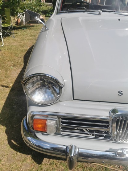The front right hand corner of a pale grey car. The car has a chrome bumper and a grille, the indicator and side light are on the edge of the grille. The car has a round headlamp, part of the bonnet line is visible.