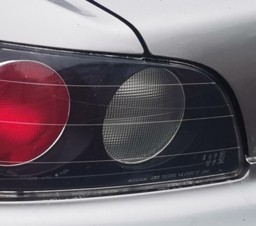 Part of the left hand rear light cluster of silver car. The lights are round and set into a black surround. The edge of the boot/trunk is visible and curves  around the edge of the light cluster.