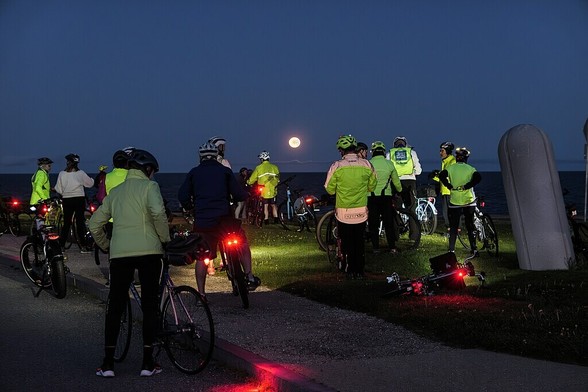 WikiCommons color photo shows approximately 15 bicyclists in full cold riding gear, from behind. They are at a waterfront observing the full moon in the distance. Photo by Kenneth C. Zirkel, 2023.

Caption: "Bike Newport's Full Moon Ride stops at Brenton Point to admire the moon."

https://commons.wikimedia.org/wiki/File:Bike_Newport_Full_Moon_Ride_at_Brenton_Point.jpg