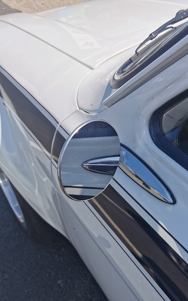 View of part of the passanger side of a white car with brown./black stripe.
The door mirror is circular and chrome.
Part of the front wing is visible with flared wheel arch and shiny wheel.