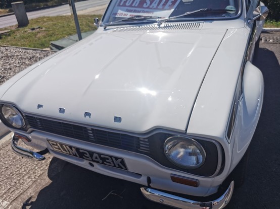 Slightly off-set view of the front of a white Ford Escort MK1 Mexico. The word Ford is visible across the front edge of the bonnet. Cut away chrome bumpers, a flared front wheel arch and signature stripe are partially visible.