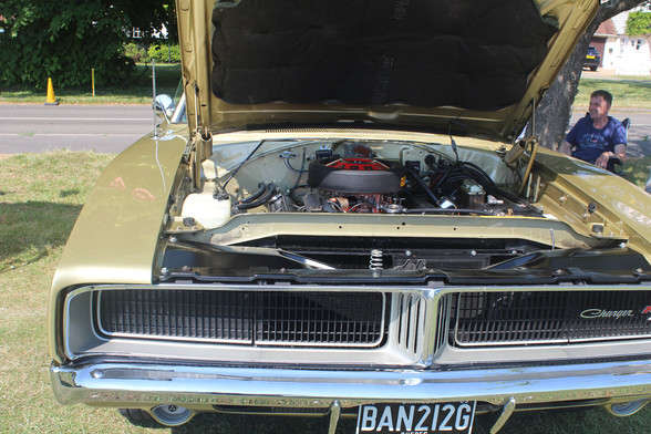 View of teh front of a late 60's Dodge Charger. The bonnet is open revealing the engine, the large carburettor is clearly visible. 
Te Charger nameplate is visible on the left-hand side of the vehicle, on the front grille.  The car is a metallic gold colour.
