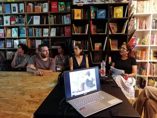Fotografia da livraria Tigre de Papel com estantes nas paredes e uma mesa grande no centro rodeada de pessoas. Na frente da imagem, vê-se um computador portátel em cima da mesa, com um desenho de origem japonesa.