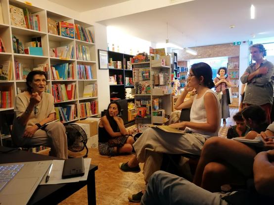 Fotografia da livraria Tigre de Papel com estantes nas paredes. Algumas pessas estão sentadas em cadeira, outras no chão, outras em pé.