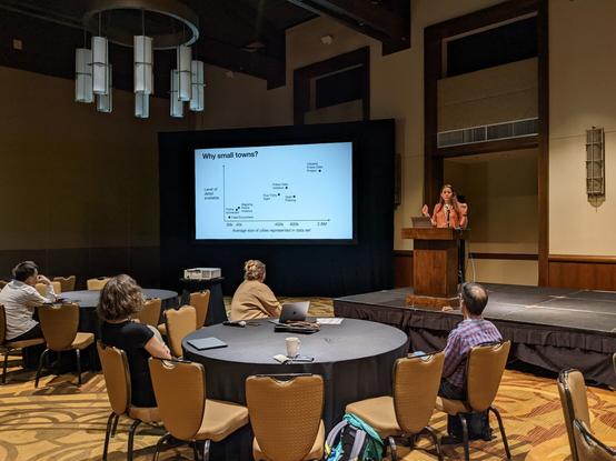 Ariana Mendible stands behind a lectern, presenting slides to a ballroom of attendees
