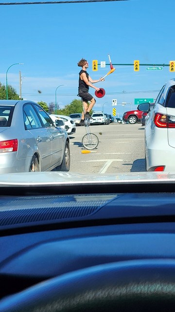 A man in shorts juggles pins and a hat while riding a unicycle at a street intersection during a red light in Vancouver.
