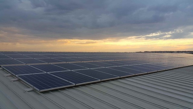 A few sunbeams peak out from a heavily clouded sky above a warehouse roof with rows of solar panels.