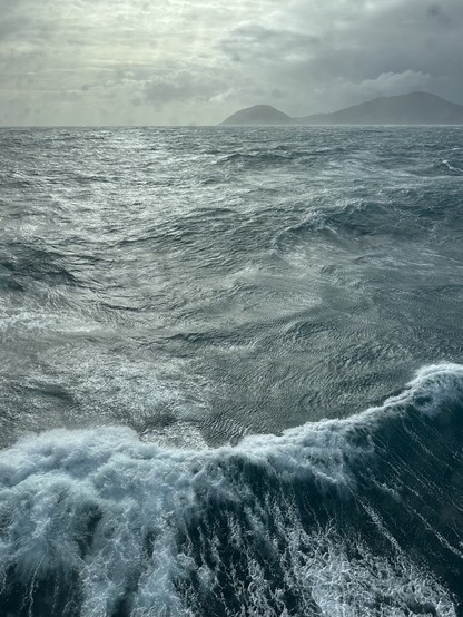 A stormy ocean view from the bridge of a research vessel with land in the far distance and a breaking wave in the foreground.