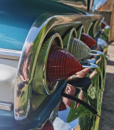 The rear light cluster of a period American car. The lights are arranged as three individual lamps each with a chrome surround. The lamps are very pointy. There is a big chrome bumper below the lights and thick chrome strip around the rear panel. A little piece of bodywork is visible it is mainly marine blue with a white stripe. There are chrome strips either side of the white stripe. The car appears to have a boot/trunk.