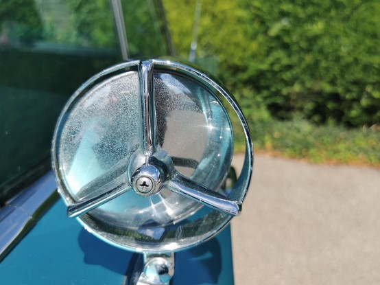The door mirror on a car with blue bodywork, viewed from the back of the mirror. The mirror has a distinctive chrome three armed support that holds the mirror glass in place.